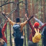 Four friends with backpacks raising arms in forest.