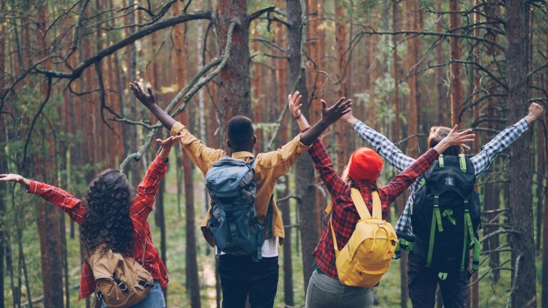 Four friends with backpacks raising arms in forest.