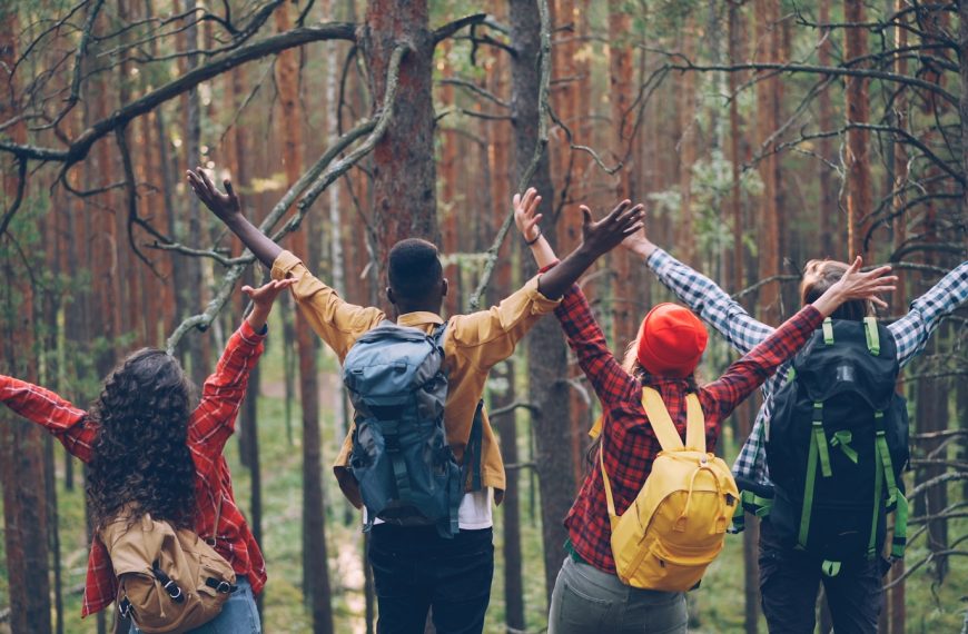 Four friends with backpacks raising arms in forest.